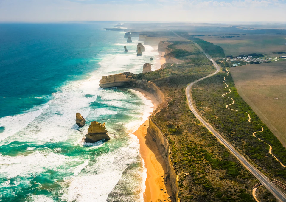 Coastal cliffs with rock formations rise from the ocean, lined by a winding road and adjacent fields extending into the distance.