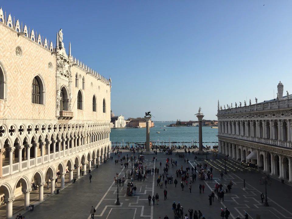 A crowded square beside a historic building with arched windows, leading to a waterfront with two statues and a view of distant islands.