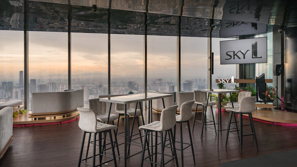 Modern rooftop lounge with high tables and chairs, large glass windows showcasing a cityscape at sunset.