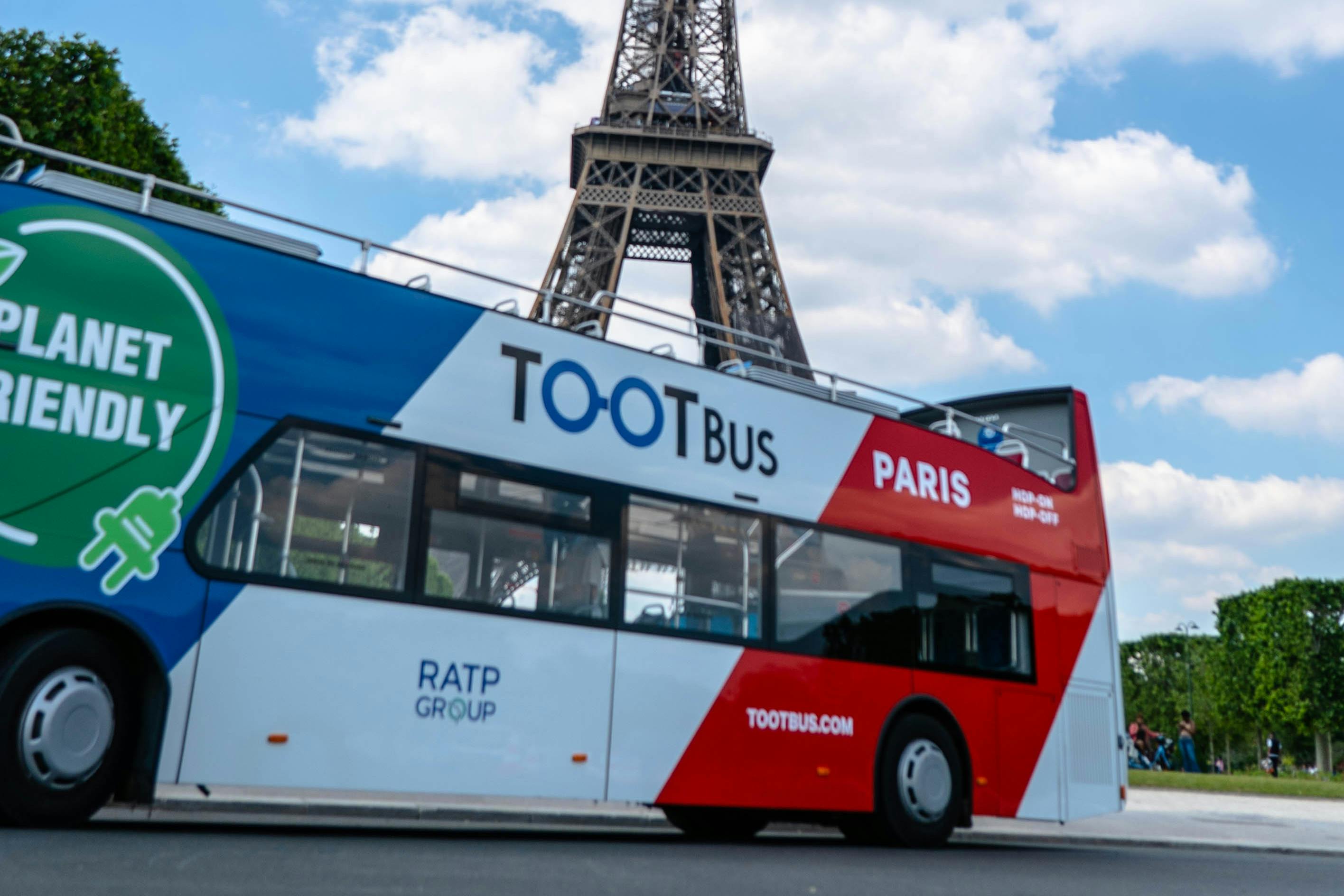 Un autobús turístic de dos pisos amb l'etiqueta "TOOTBus Paris" amb la Torre Eiffel al fons contra un cel parcialment ennuvolat.