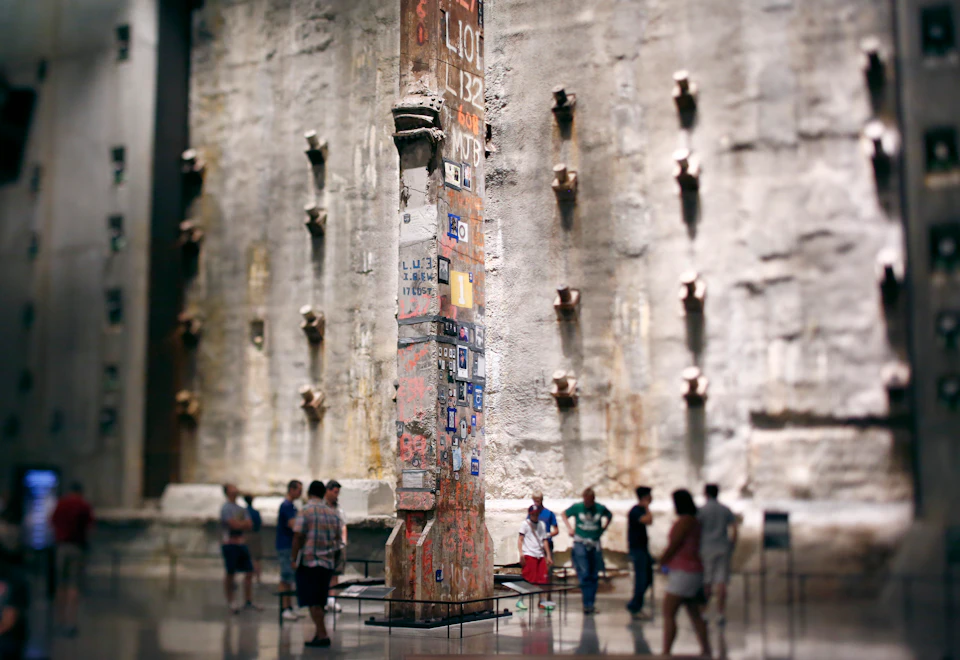 People observe a tall, decorated steel column in a large, industrial-looking room with rugged, gray concrete walls.