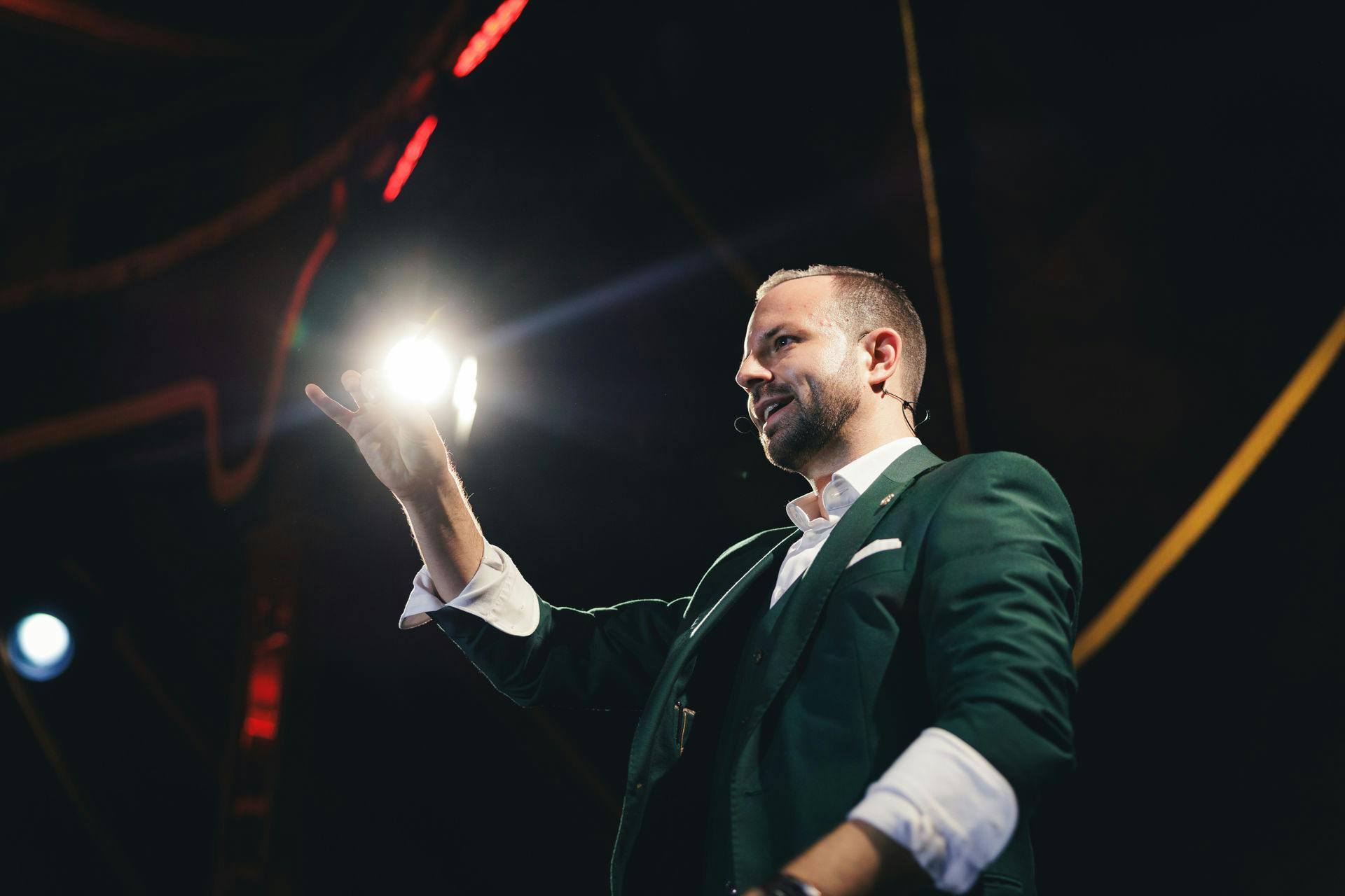 一名身穿绿色西装的男子在舞台上用麦克风耳机说话，在黑暗的环境中，聚光灯照亮了他。
