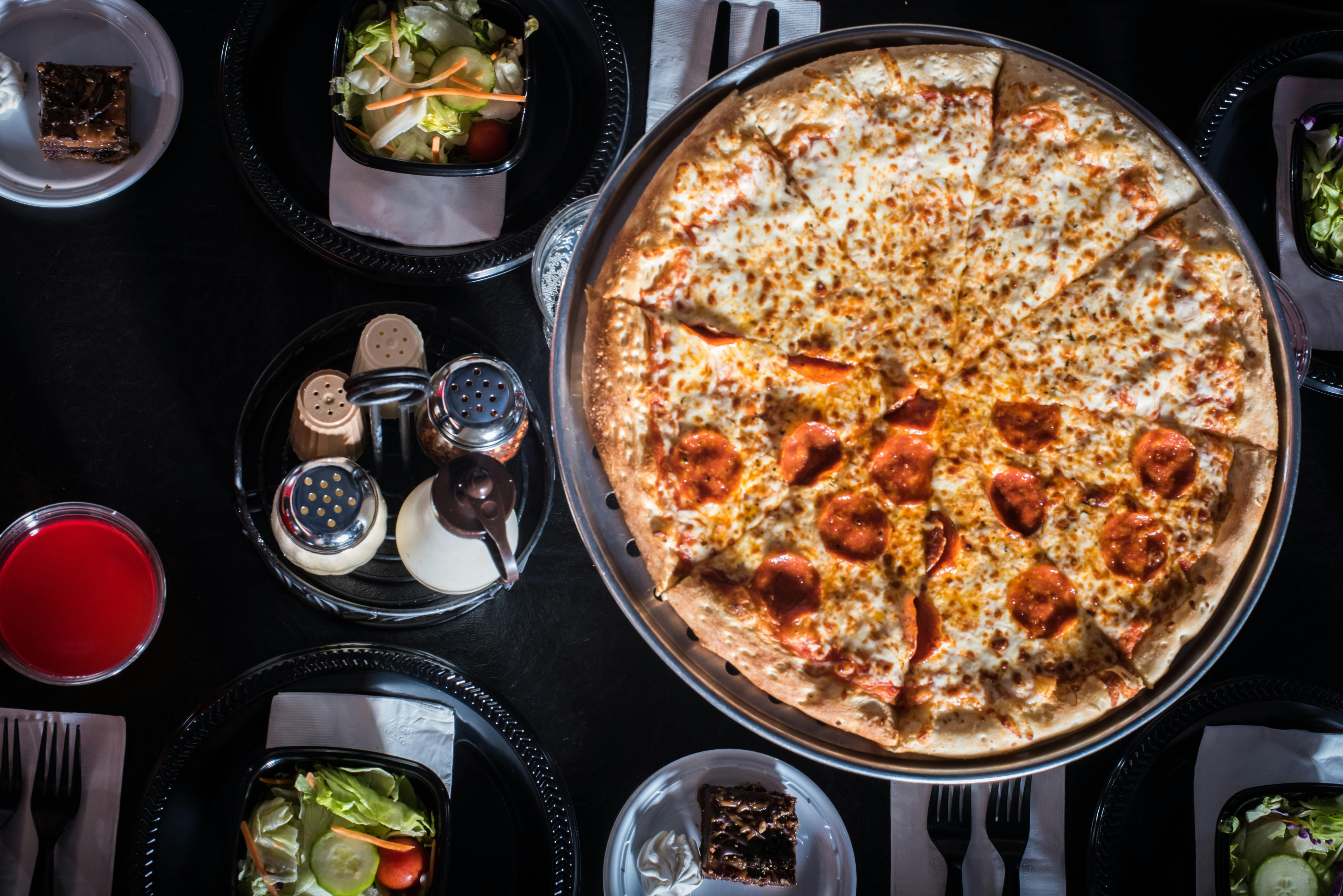 大意大利香肠披萨，配两份沙拉、盐、胡椒、奶酪摇动器、调料和黑色桌子上的甜点布朗尼。