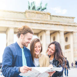 Tre personer som ler och tittar på en karta, står framför en stor historisk port med pelare och en staty ovanpå.