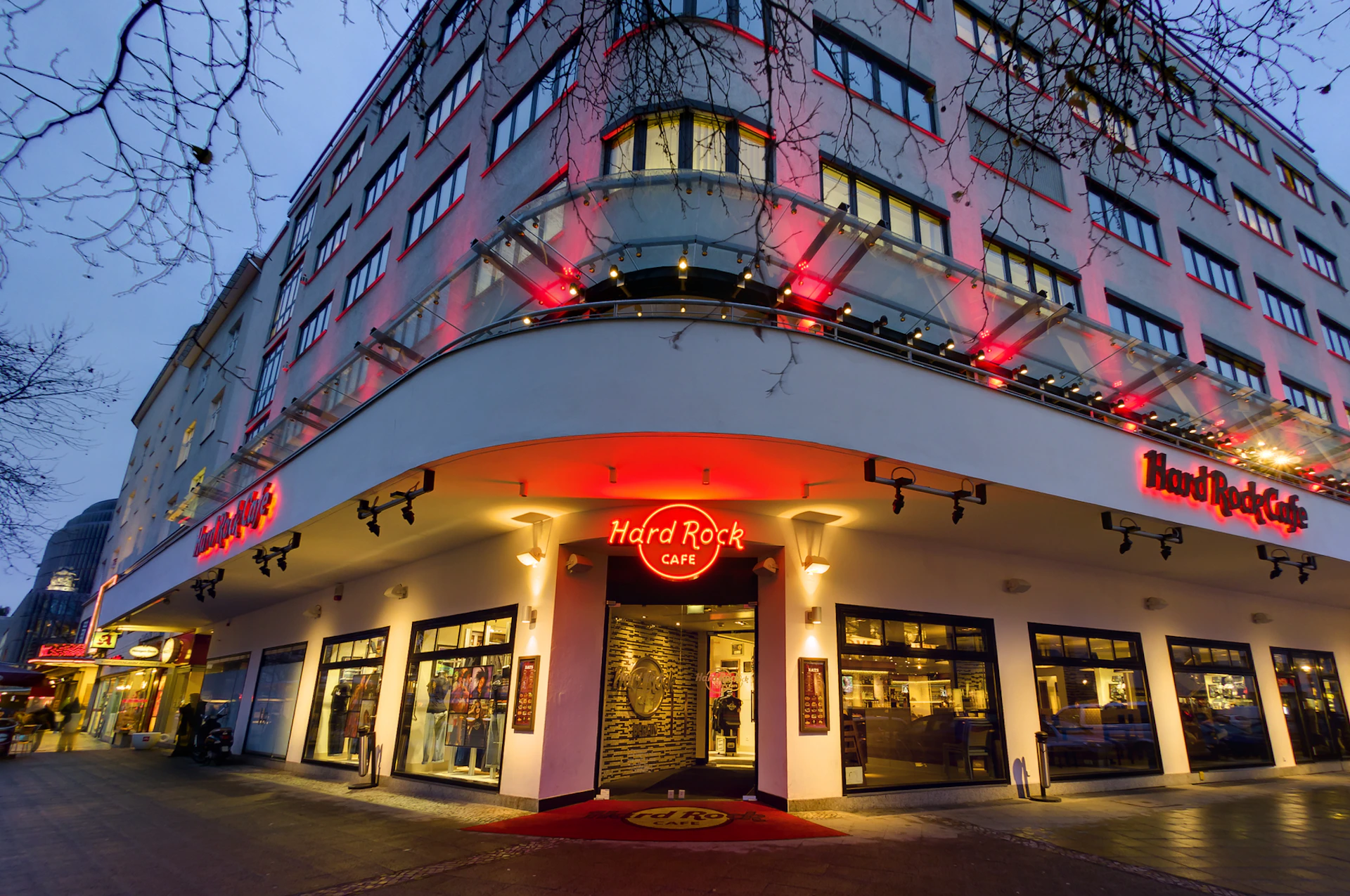 Storefront of a Hard Rock Café with illuminated red sign and entrance lights, set in a multi-story building at dusk.