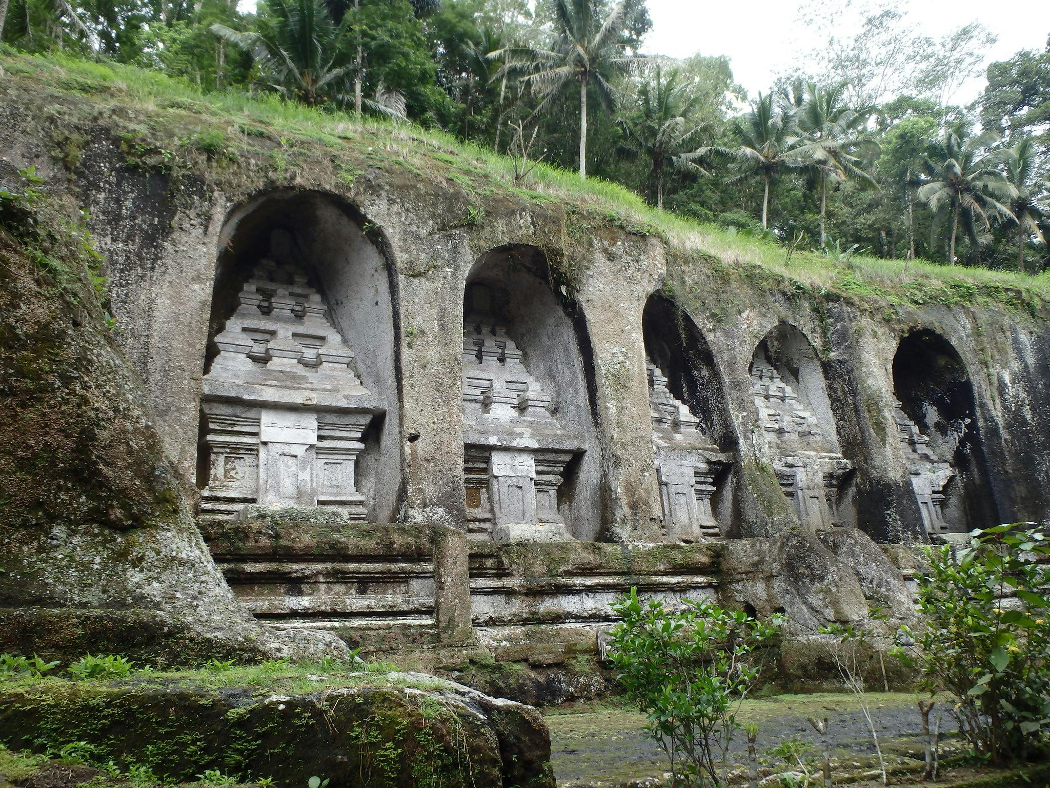 Stone carvings of temple shrines embedded into a rocky hillside with surrounding greenery and palm trees.