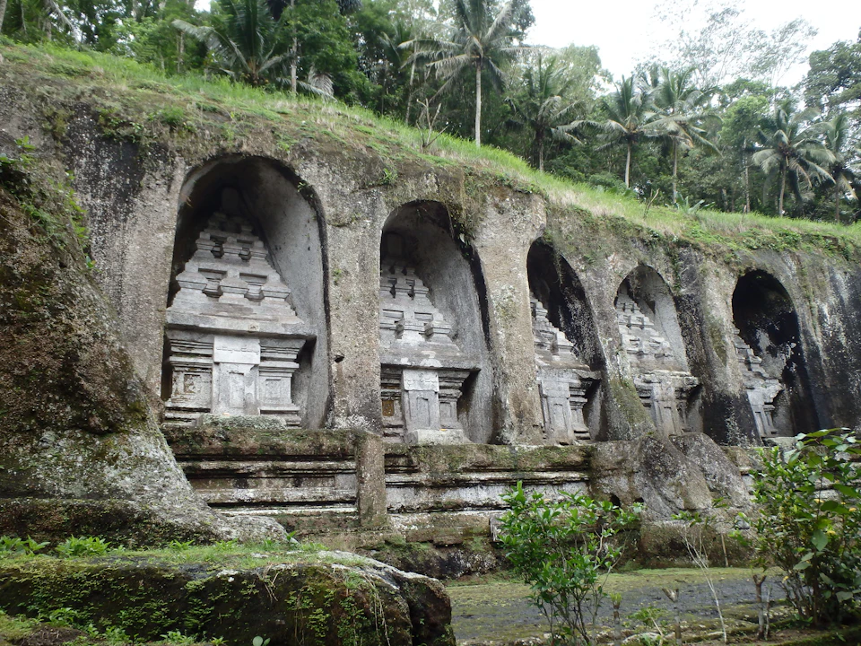 Stone carvings of temple shrines embedded into a rocky hillside with surrounding greenery and palm trees.