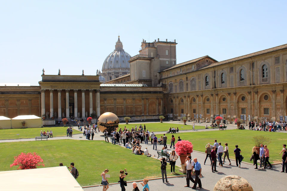 A courtyard with green grass, people walking, a bronze sculpture, and historical buildings under a clear blue sky.