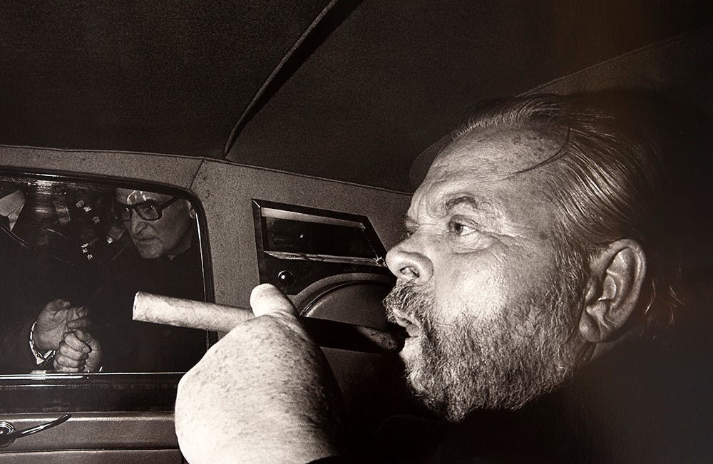 Elderly man with a thick beard, holding a cigar, close-up profile view inside a dimly lit space.