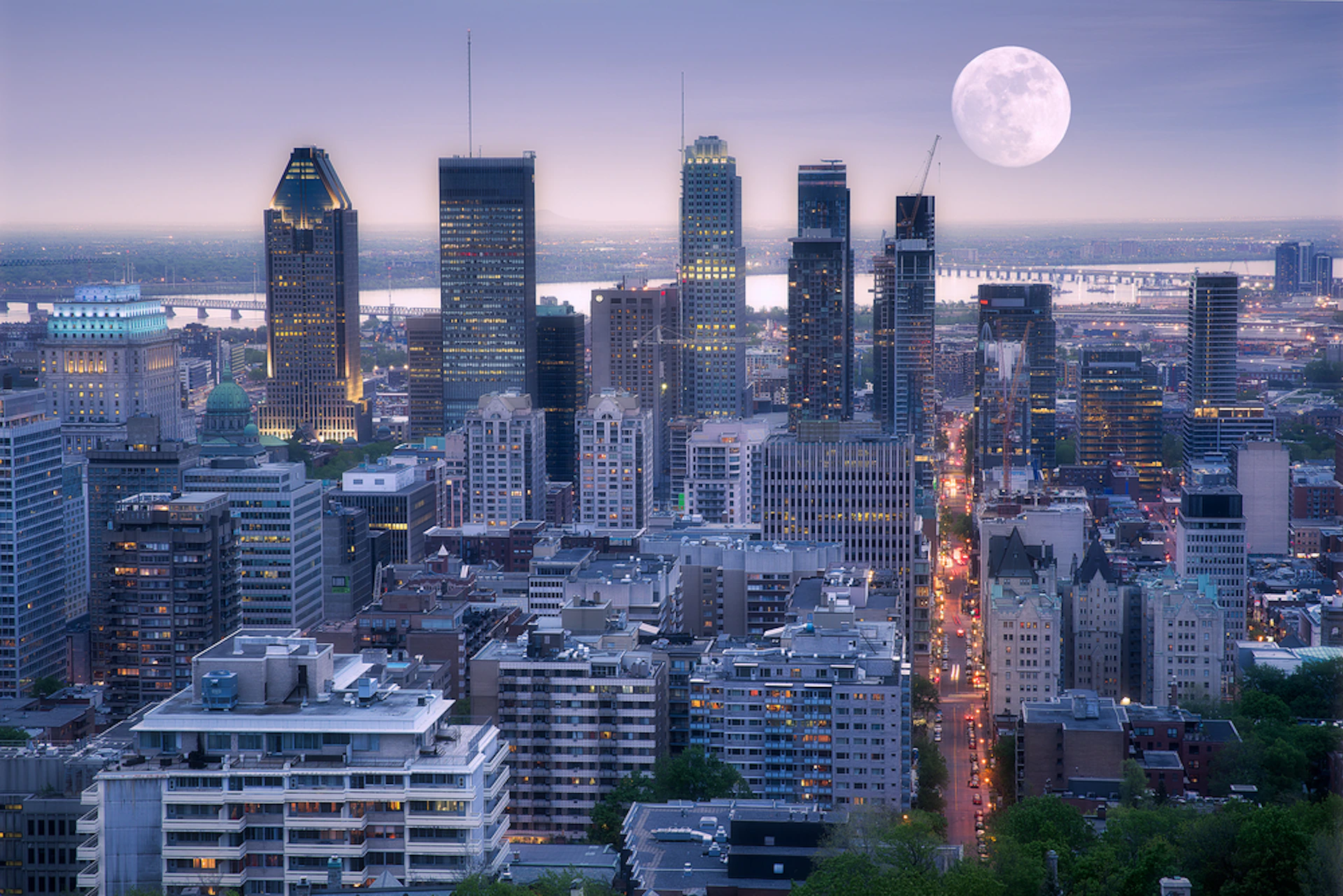 City skyline at dusk with tall buildings and a full moon rising. Lights illuminate the streets and buildings.