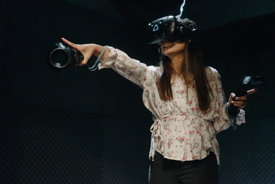 A person wearing a floral blouse uses a virtual reality headset and controller, extending one arm forward, smiling.