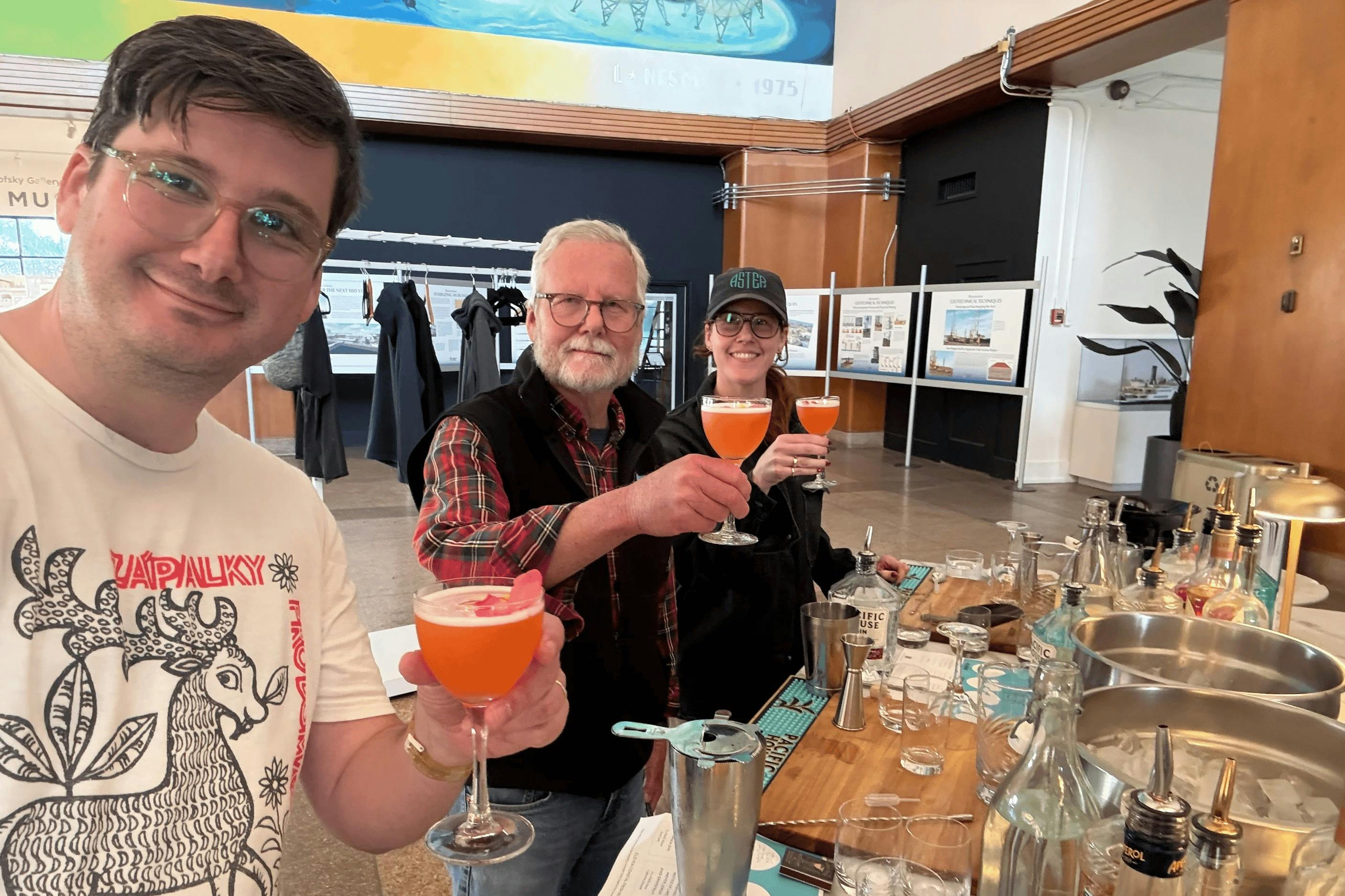 Three people smiling and holding orange-colored drinks in a social setting with a museum-like interior in the background.