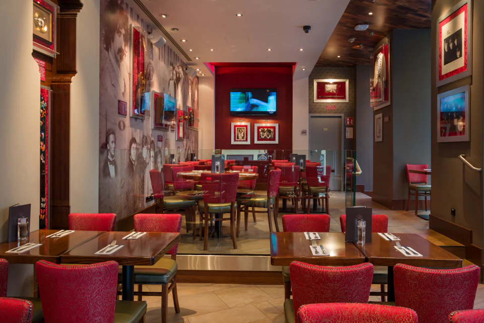 Restaurant interior with red chairs, wooden tables set with cutlery, wall art, and a TV screen on a red accent wall.