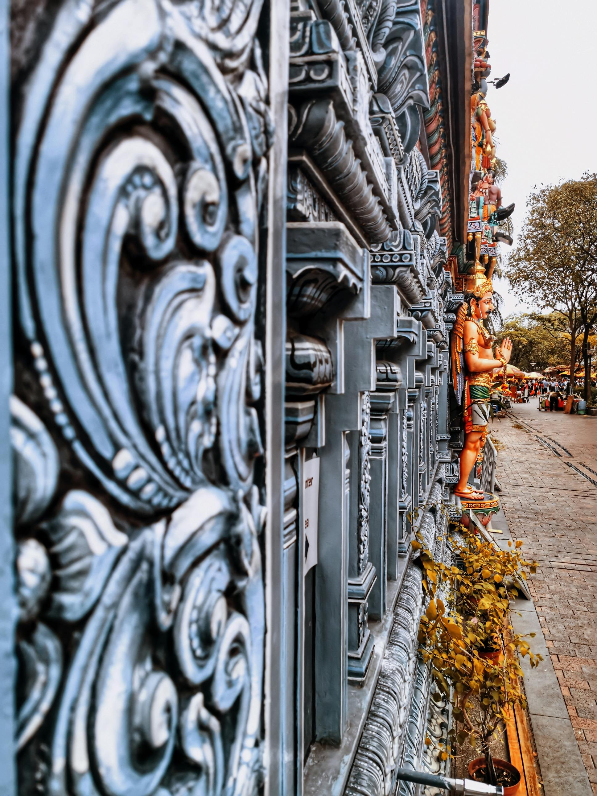 Close-up of an intricately carved temple wall, with colorful statues and trees lining a pathway in the background.