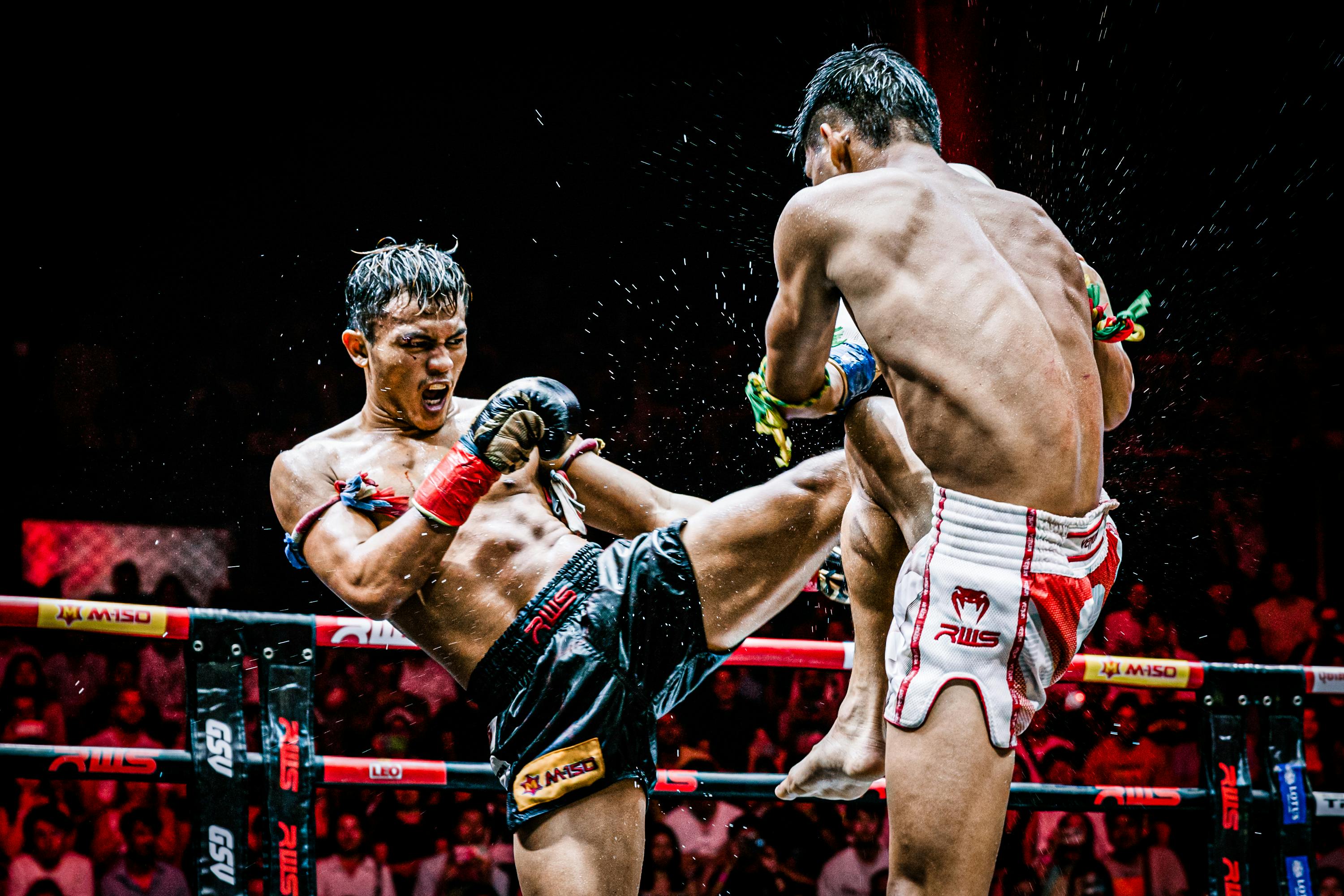 Two fighters in a boxing ring during a match, one delivering a kick to the other. Crowd visible in the background.