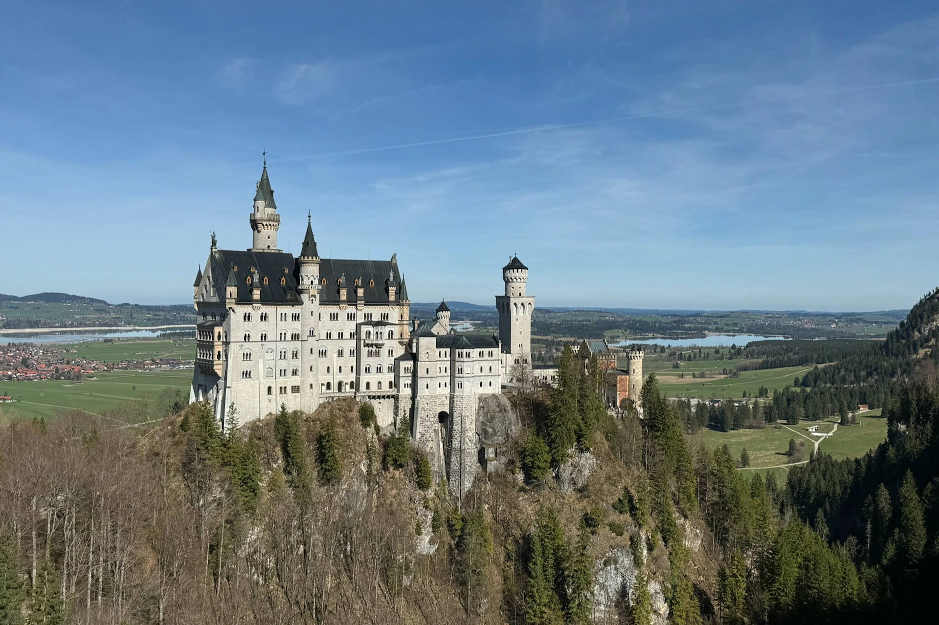 A castle with turrets and towers on a rocky hilltop surrounded by trees, with a view of fields and a lake in the distance.