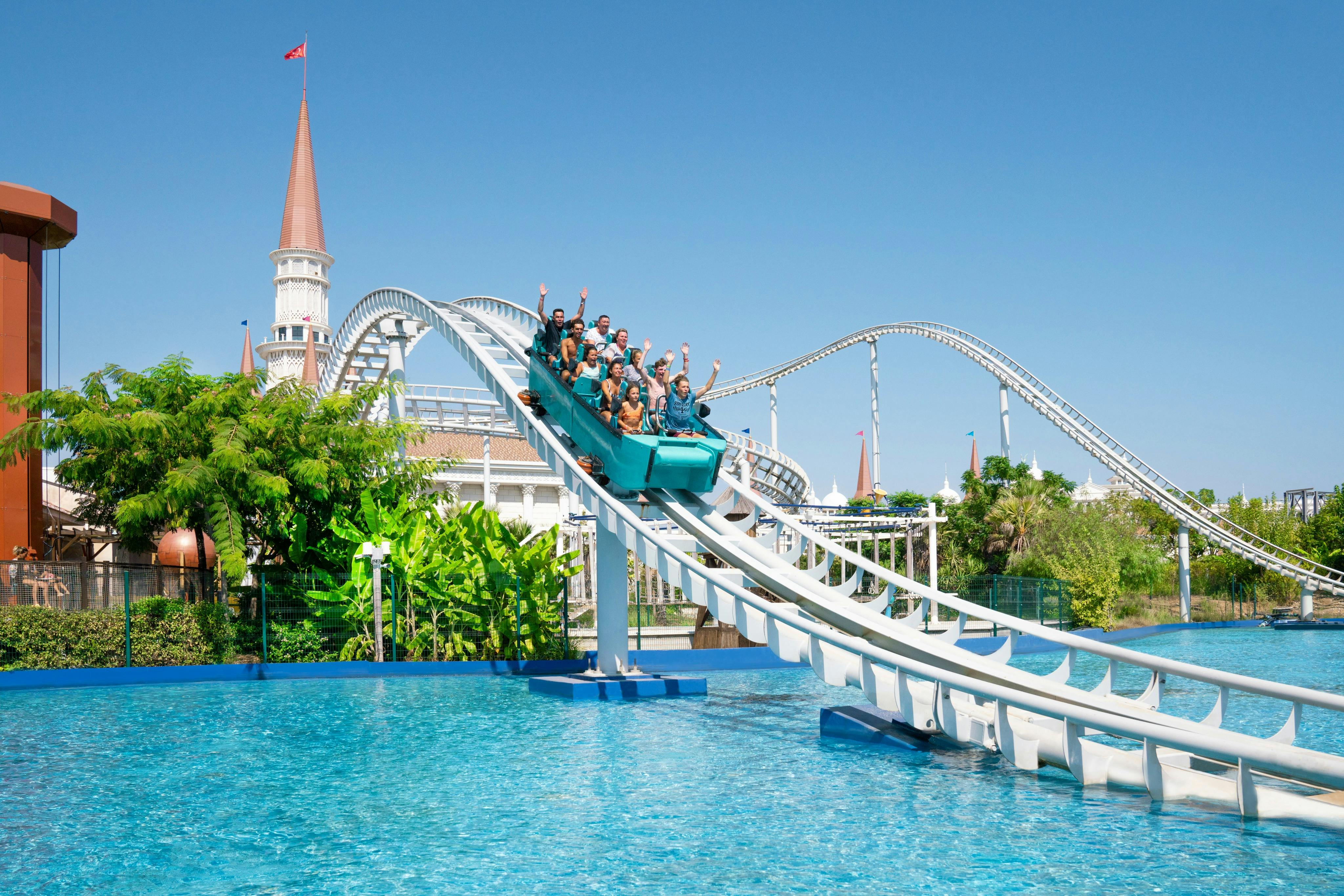 Un groupe de personnes chevauchant des montagnes russes au-dessus de l'eau dans un parc d'attractions, avec un ciel bleu et une structure ressemblant à un château à l'arrière-plan.
