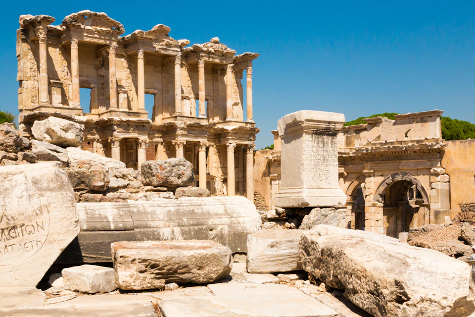 Ancient ruins with eroded columns and stone blocks under a clear blue sky. Partially standing structure in the background.