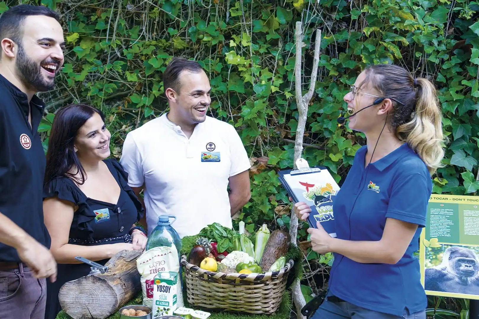 Four people stand outside near greenery, smiling and talking. A basket of fruits, vegetables, and groceries is on a table between them.