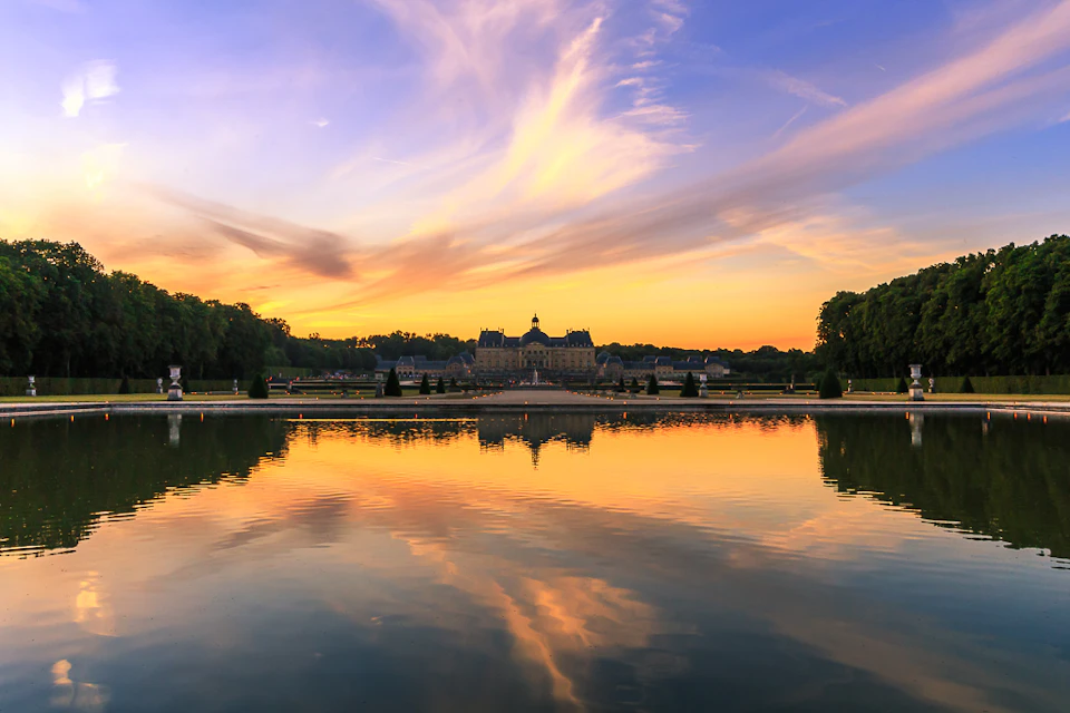 A grand building at sunset, reflecting in a calm pond with vibrant clouds and silhouettes of surrounding trees.