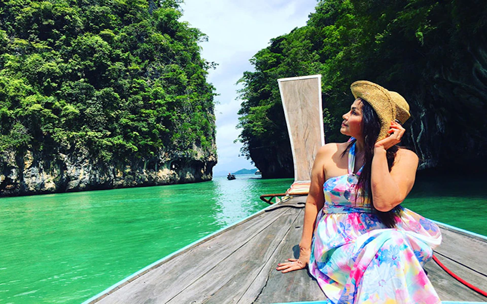 A woman in a colorful dress and straw hat sits on a boat in a turquoise waterway surrounded by lush green cliffs.