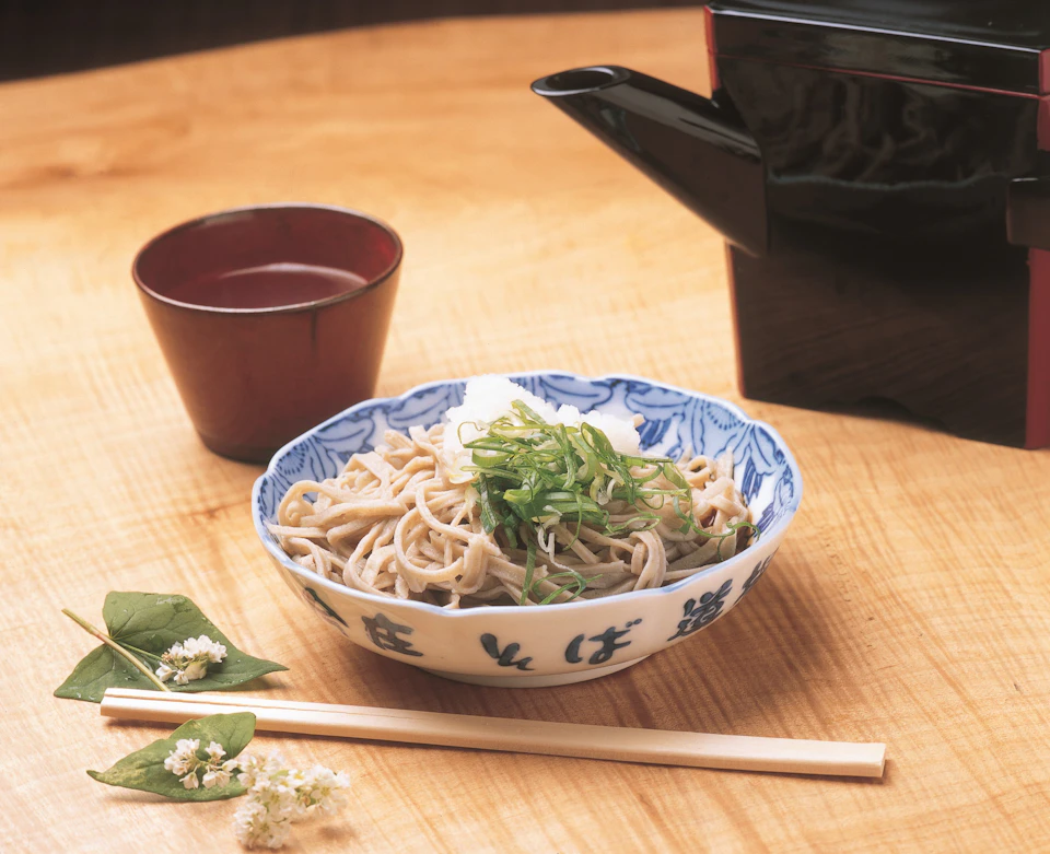 A bowl of soba noodles with garnish, a cup, chopsticks, and a black teapot on a wooden table with a floral accent.