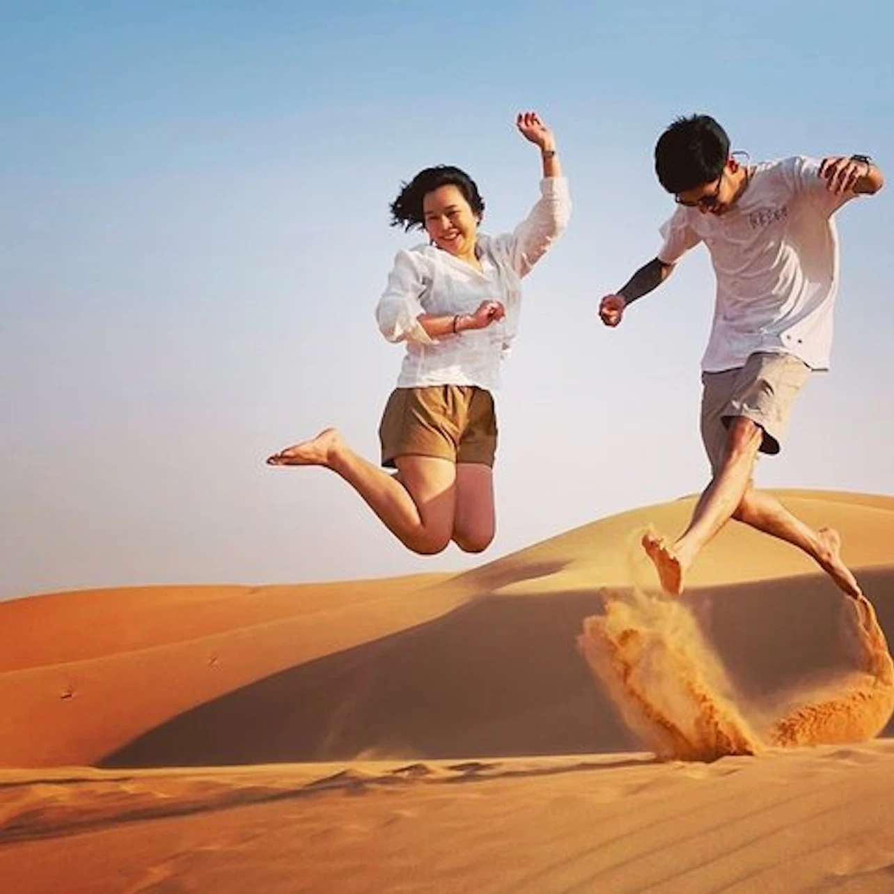 Two people jump joyfully on sand dunes under a clear blue sky; one kicks up sand with his foot.