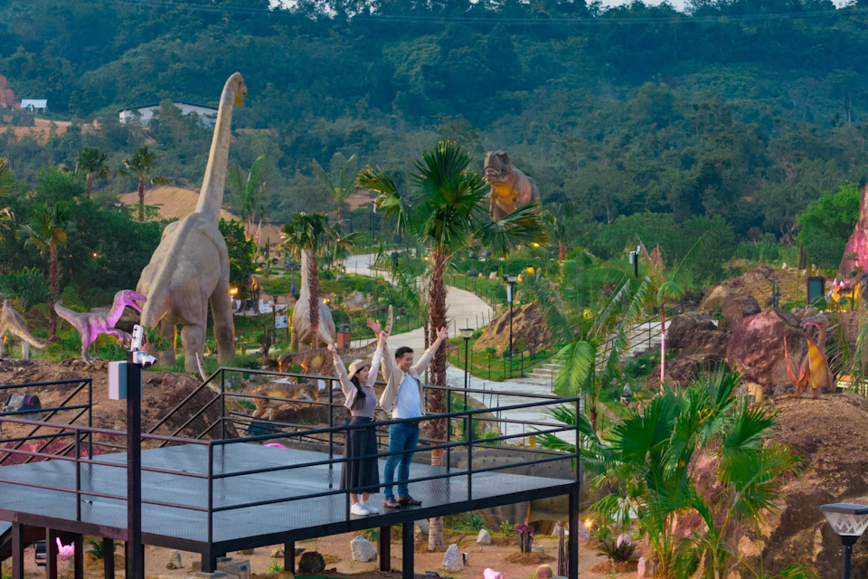 Two people on a platform in a park with life-size dinosaur statues and lush greenery.