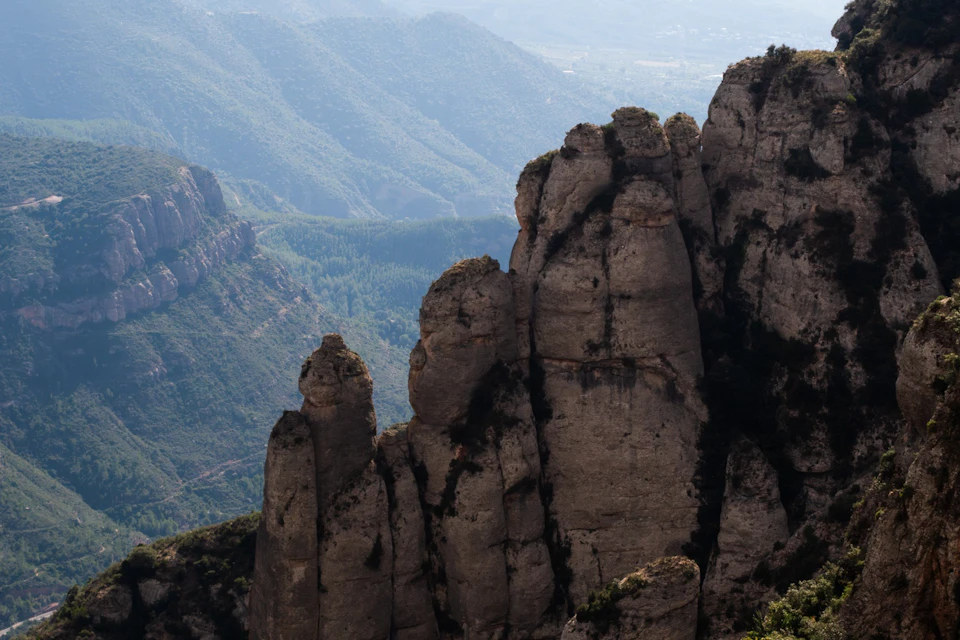 Tall rocky peaks with rugged surfaces rise against a backdrop of forested hills and distant hazy mountains.