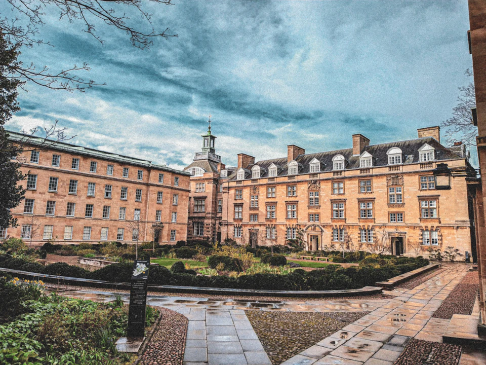 Brick building complex with a central courtyard and pathways, surrounded by hedges. Cloudy sky in the background.