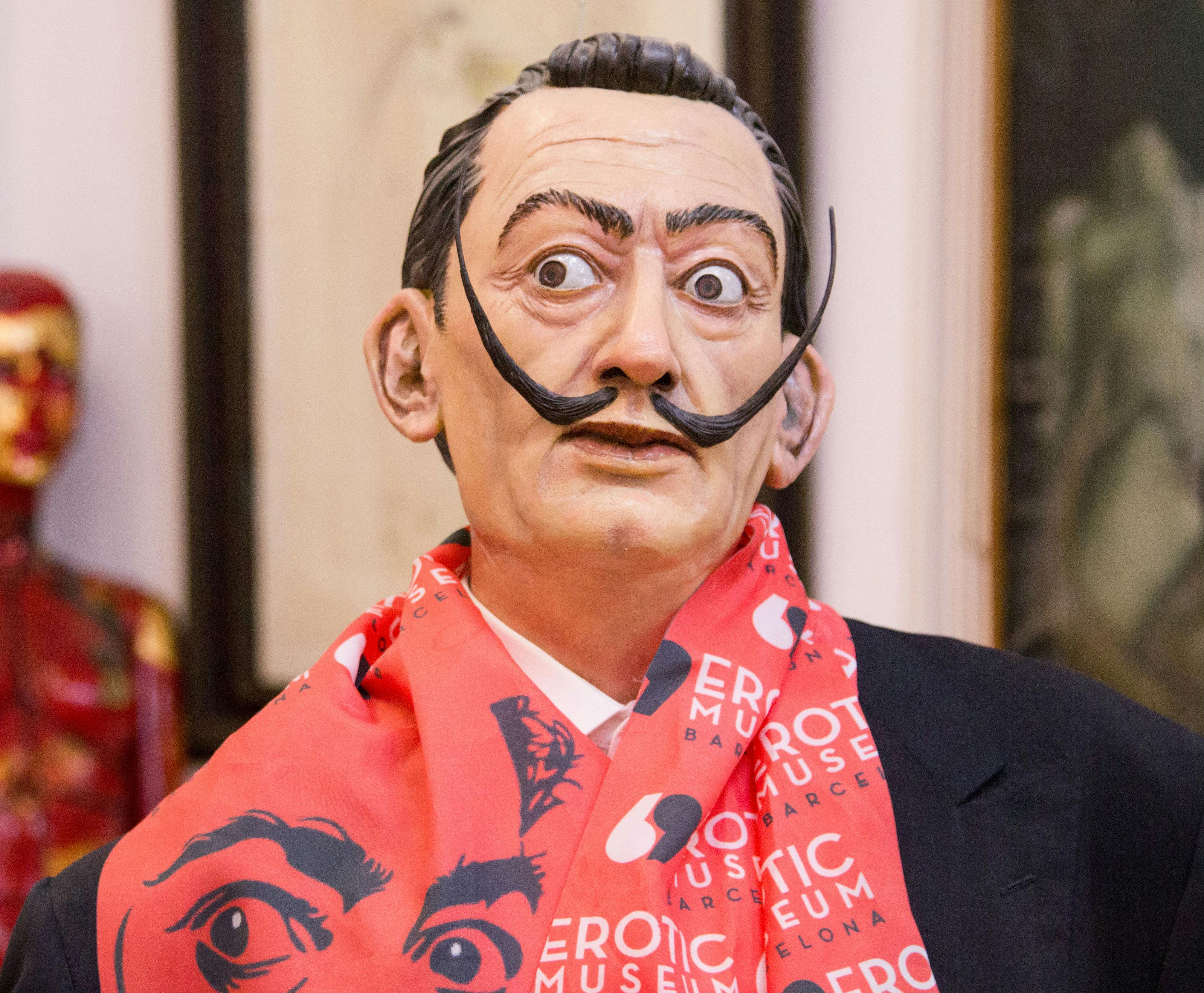 Živoucí socha muže s výrazným knírem, červenou šálou s textem „Erotické muzeum Barcelona“ a oblekem.