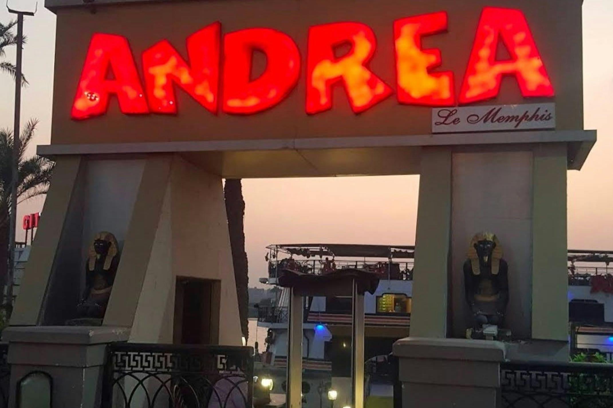Ein beleuchtetes Schild mit der Aufschrift "Andrea" über einem Eingang, mit Skulpturen und einem Blick auf eine Uferpromenade bei Sonnenuntergang.