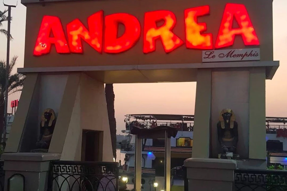 An illuminated sign reading 'Andrea' above an entrance, with sculptures and a view of a waterfront at sunset.