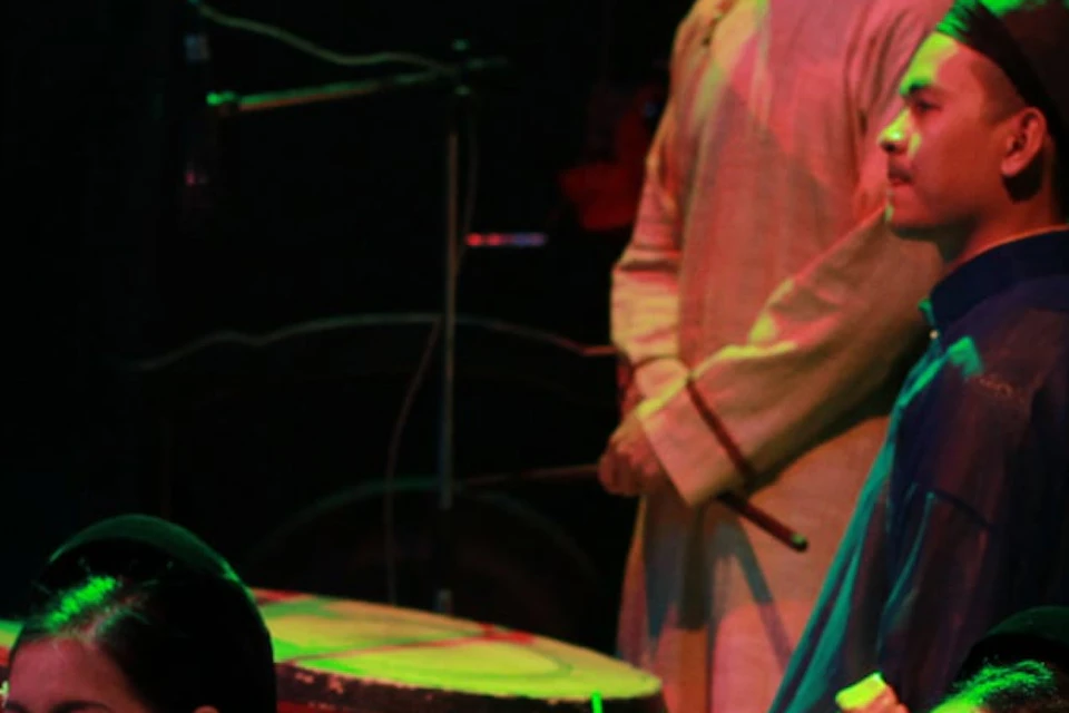 A group of performers in traditional attire playing instruments and singing on a dimly lit stage with colorful lighting.