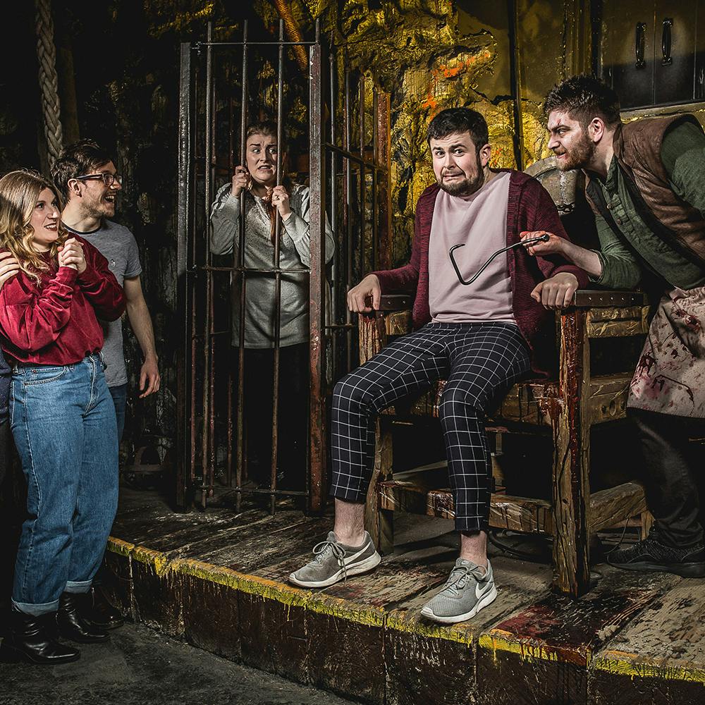 A man in a bloody apron holds a syringe near a nervous man seated in an electric chair, while others watch and a person is locked in a cage.