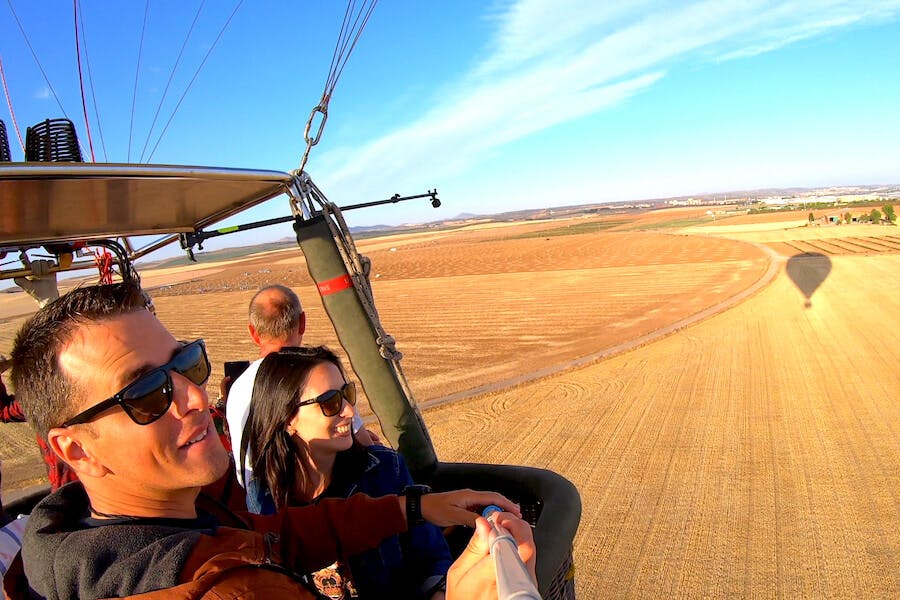 三个人乘坐热气球自拍，背景是宽阔的田野和晴朗的蓝天。