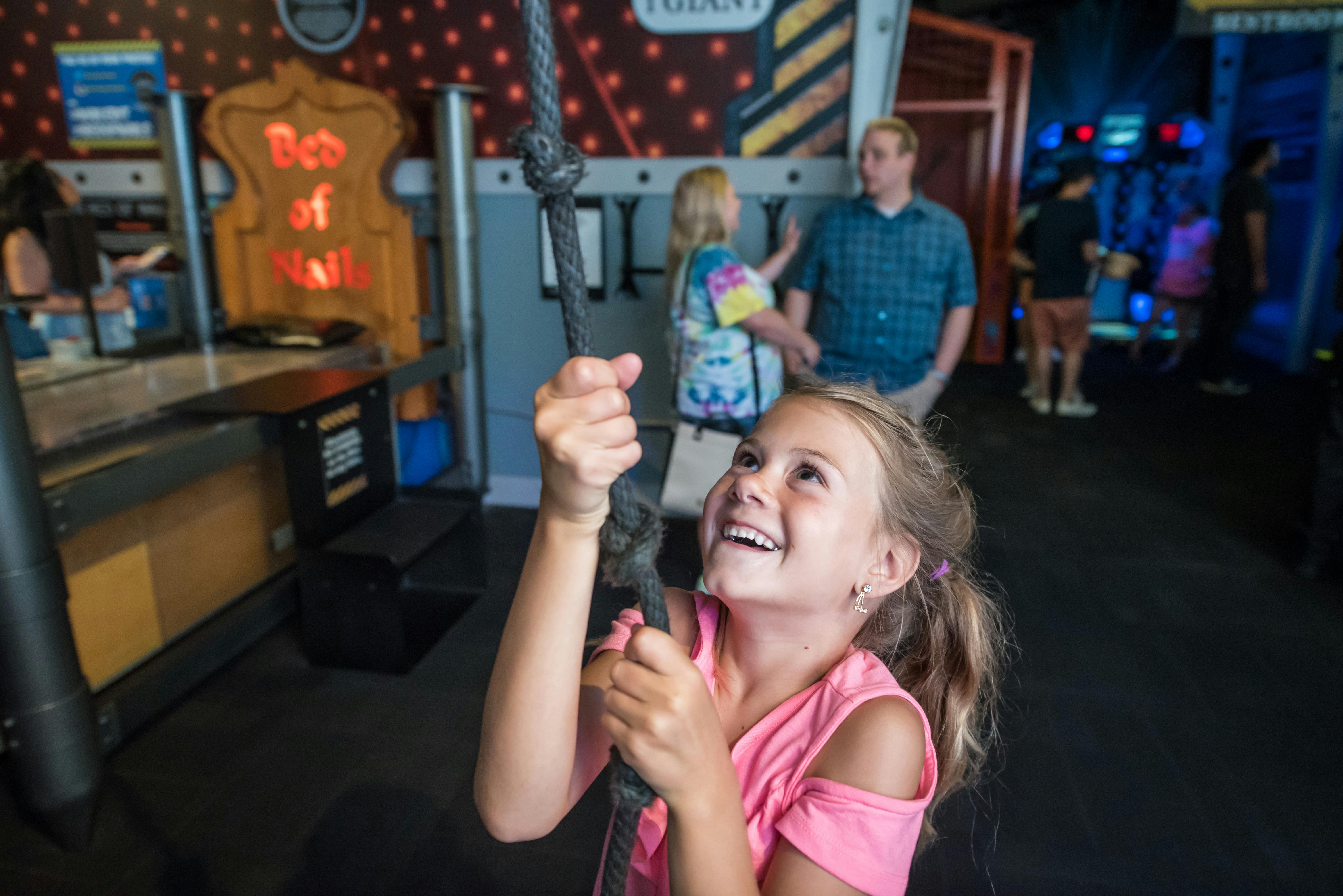 Smiling girl in a pink shirt holds a climbing rope. People, including a couple, stand in the background at an indoor activity center.