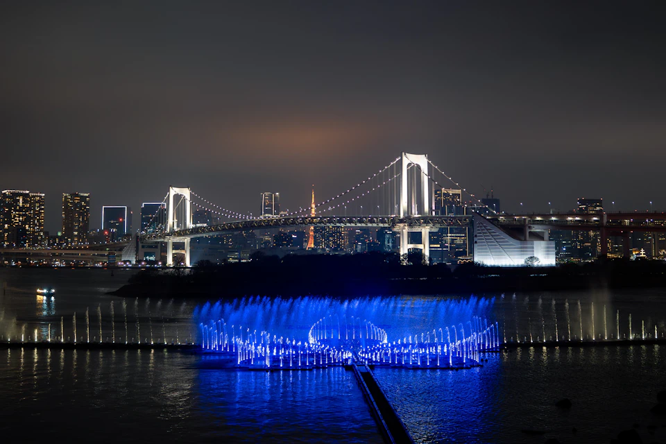 A lit-up bridge at night with a blue light fountain display on the water in the foreground.