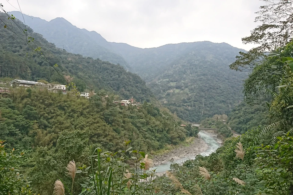 A lush green valley with small houses on the hillside, a river flowing through the center, and mountains in the background.