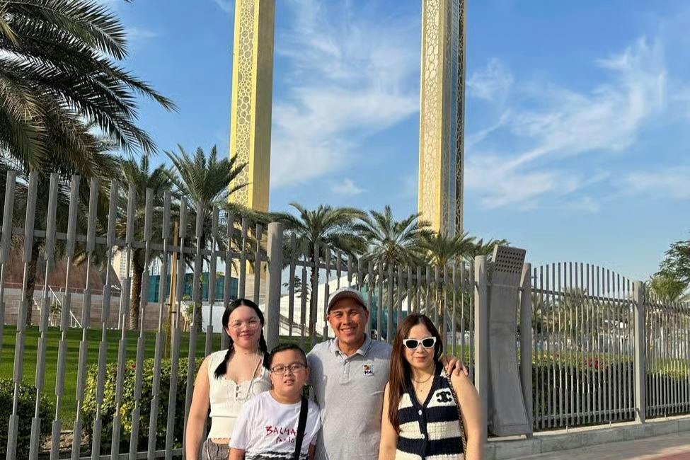 A group of four people stands in front of a decorative golden rectangular structure with palm trees and a fence in the background.