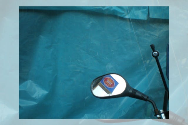 A motorcycle mirror reflecting a sticker with indistinct text, against a blue tarp background.