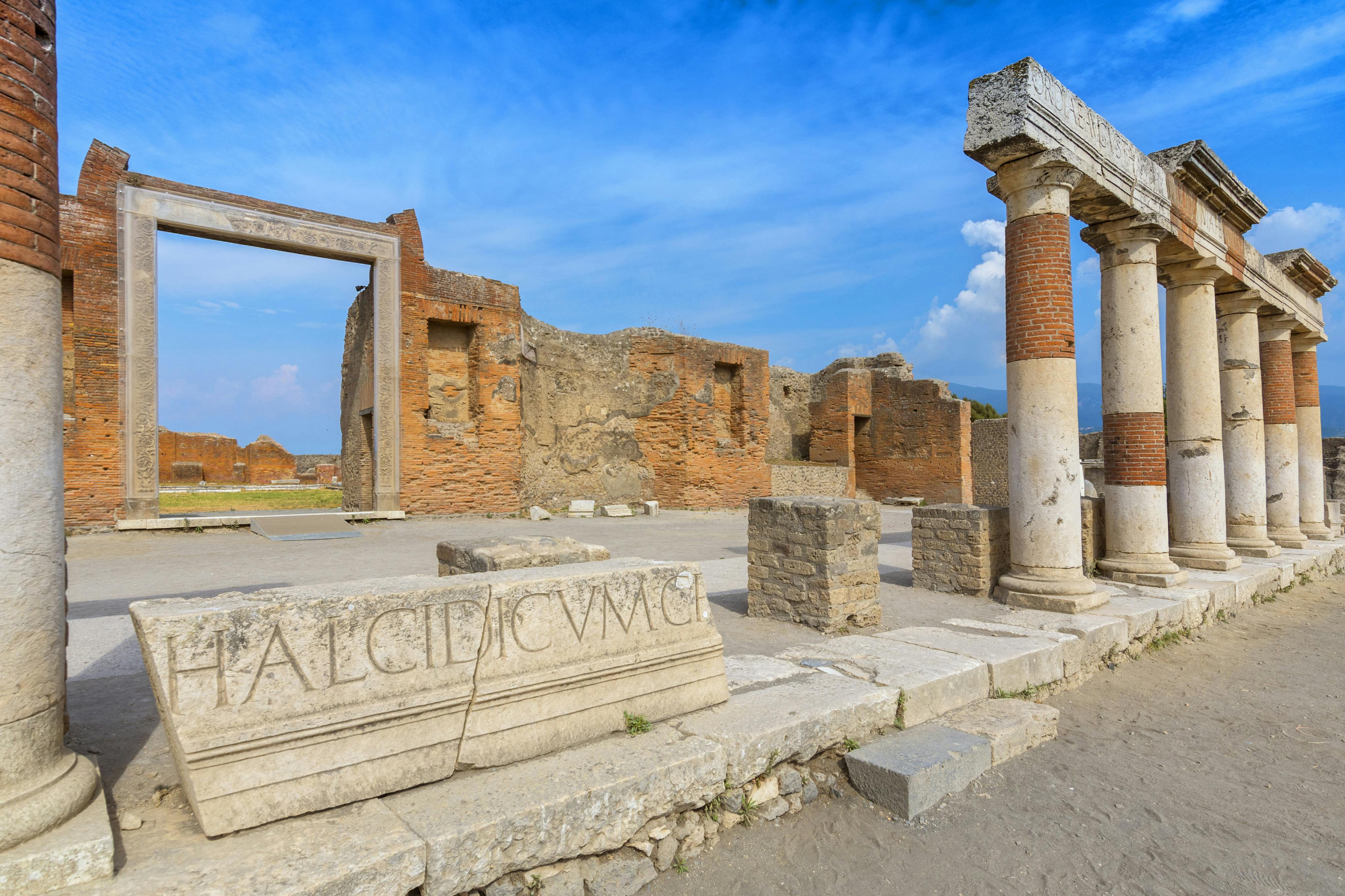 Ancient Roman ruins with partially intact brick walls, columns, and a stone block featuring an inscription, under a blue sky.