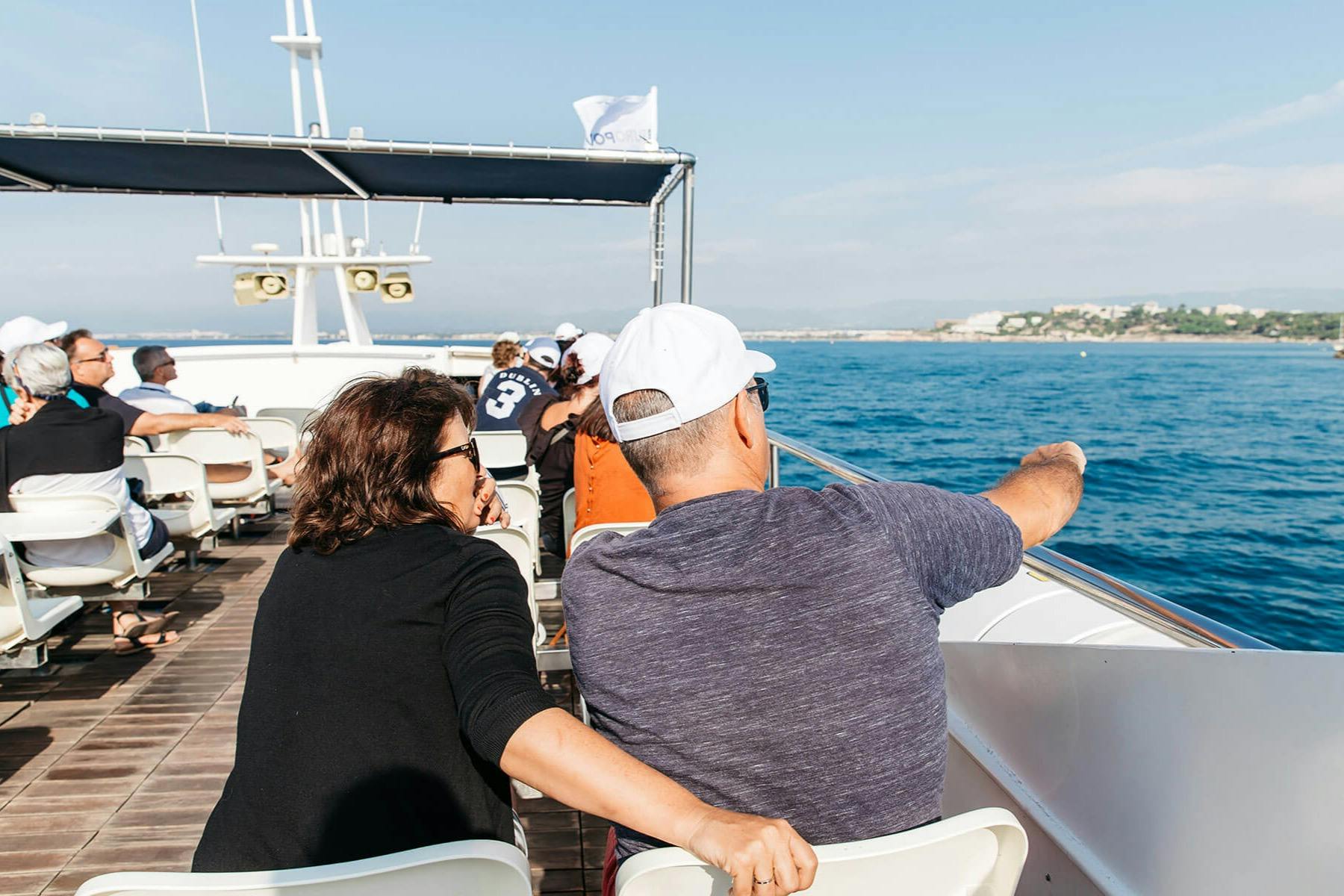 Um casal está sentado no convés de um barco, com o homem a apontar para o mar. Outros passageiros e uma linha costeira são visíveis ao fundo.