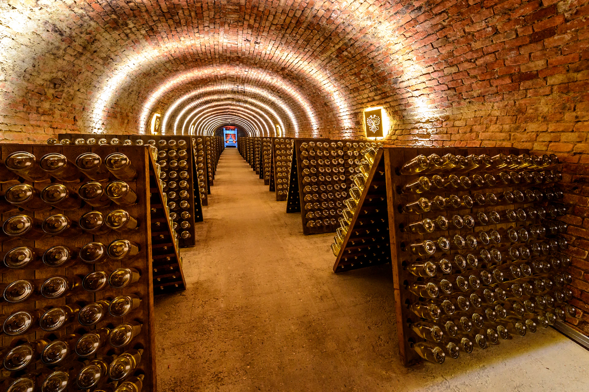 Dimly lit brick cellar with rows of wine bottles resting on angled racks, extending into the distance.