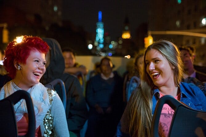 Une personne aux cheveux roux sourit alors qu'elle est assise dans un bus de tourisme nocturne avec d'autres personnes ; les lumières de la ville et les bâtiments sont à l'arrière-plan.