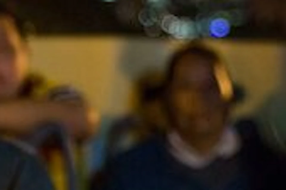 A person with red hair smiles while sitting on a night city tour bus with others; city lights and buildings are in the background.