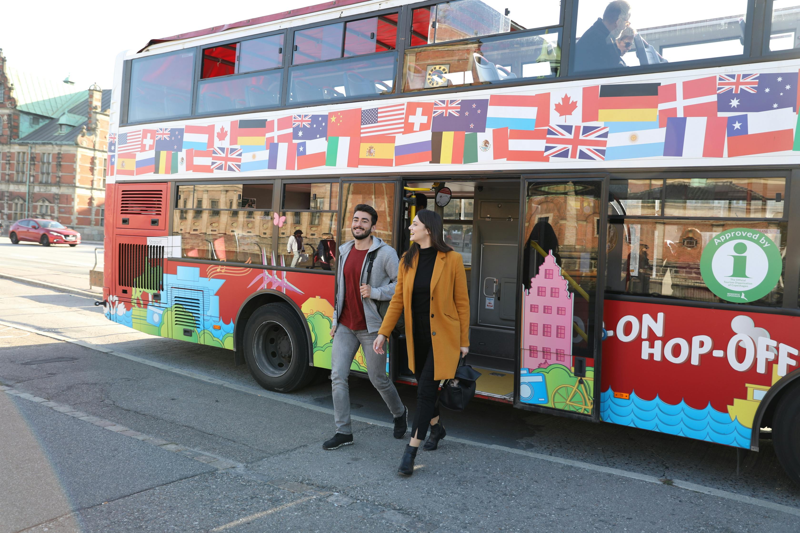 A man and woman exit a colorful double-decker bus decorated with various international flags.