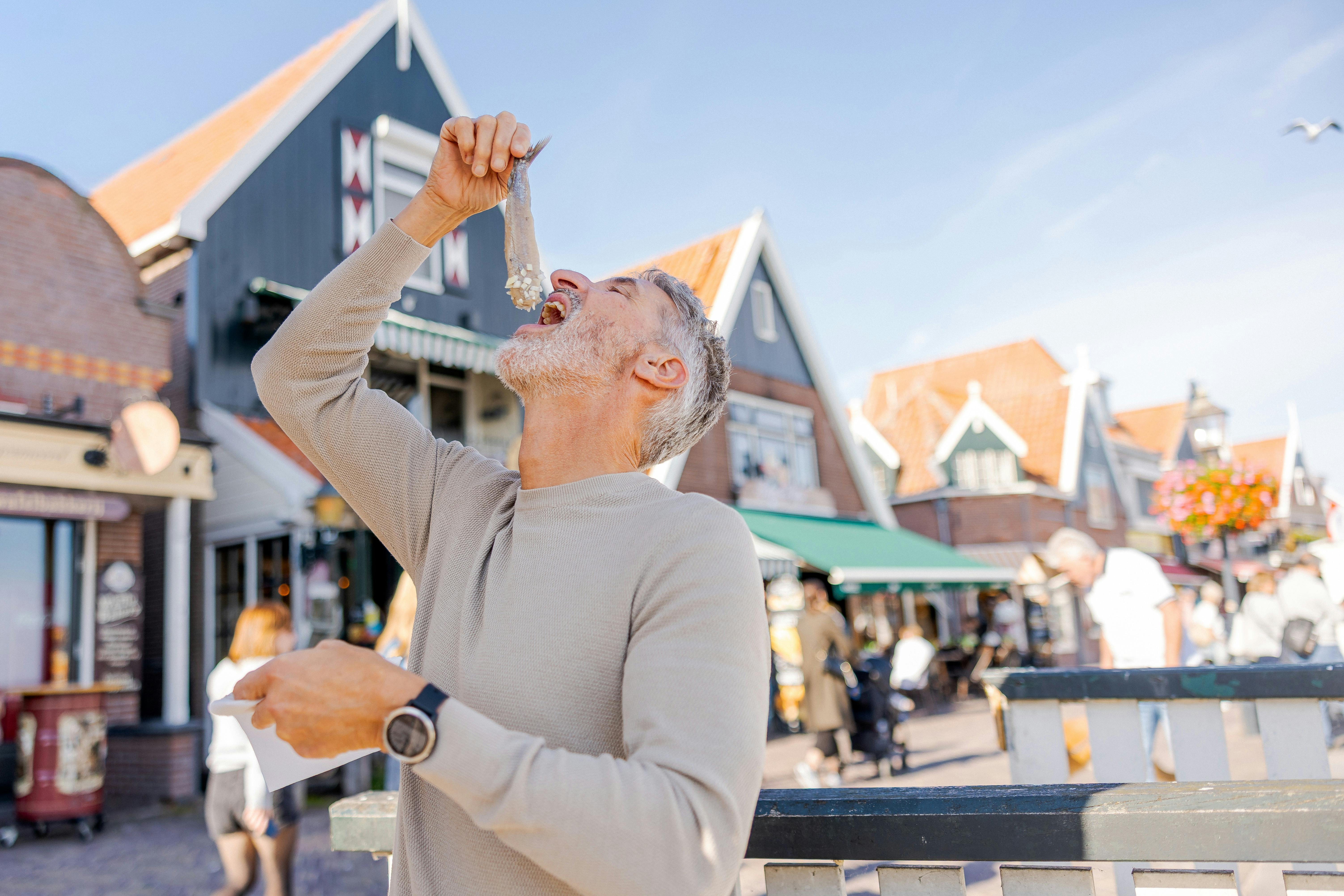 A man in a beige jumper eats a fish while standing in a market square with buildings in the background.