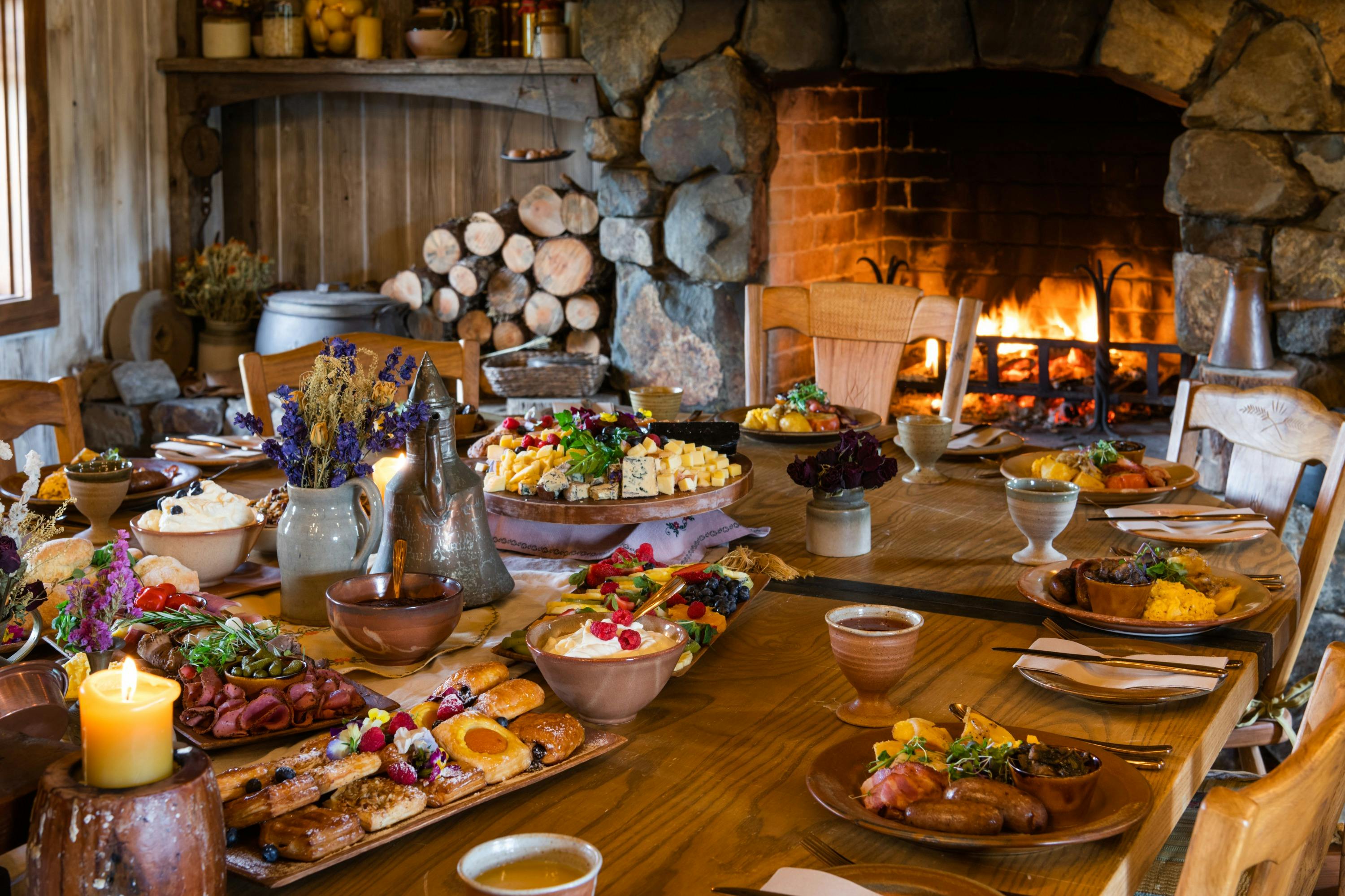 A cozy dining table set with various breakfast foods and drinks, near a lit stone fireplace in a rustic setting.