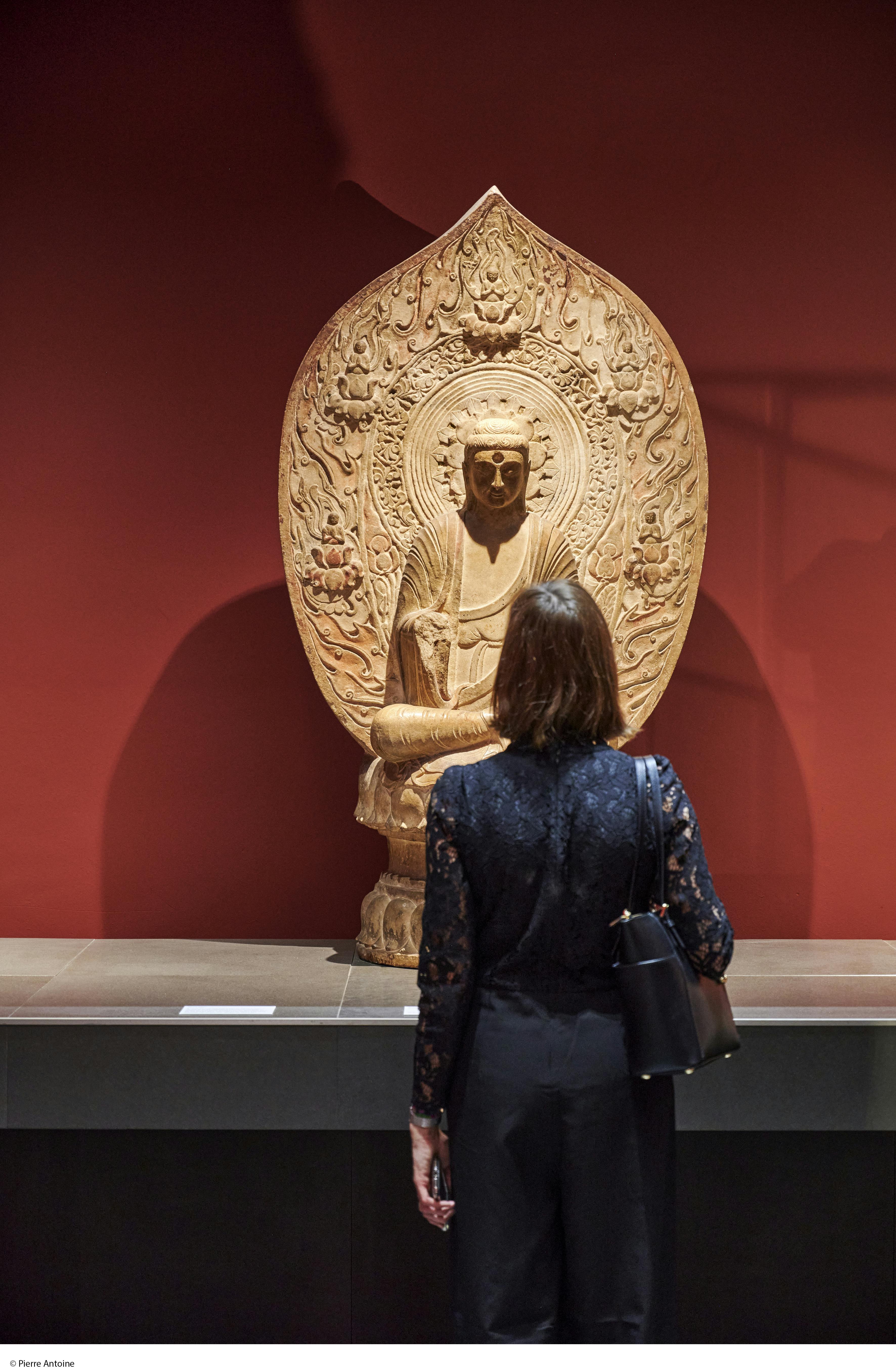 在红色背景的博物馆中，一个人站在一尊精致的佛像前。